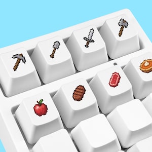 Pode incluir: Um teclado mecânico branco com teclas personalizadas impressas com ícones pixelados, incluindo uma picareta, pá, espada, machado, maçã, carne grelhada, bife cru e uma torta de abóbora dispostos em uma base branca.