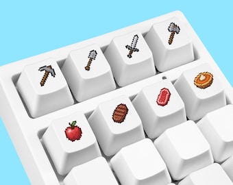 Könnte beinhalten: Eine weiße mechanische Tastatur mit individuellen Tastenkappen, die verpixelte Symbole wie eine Spitzhacke, Schaufel, Schwert, Axt, Apfel, gebratenes Steak, rohes Rindfleisch und einen Kürbiskuchen auf einer weißen Basis zeigen.