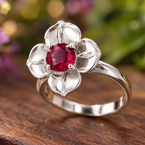 Puede incluir: Un anillo de plata con una piedra preciosa roja oscura de corte redondo engastada en el centro de un soporte de metal con forma de flor de cuatro pétalos. Los pétalos tienen líneas grabadas y la banda es de metal plateado pulido.