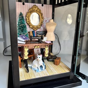 Könnte beinhalten: Ein Miniatur-Diorama in einer schwarzen Metalllaterne, das ein Nähzimmer mit einem Holztisch, einer schwarzen Nähmaschine, einer Schneiderpuppe, einem goldgerahmten Spiegel, einem kleinen grünen Baum, einer weißen Katzenfigur und einem Koffer zeigt.
