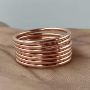 May include: A stack of seven thin, smooth, rose gold-coloured metal rings. Each ring features a simple, circular band design with a polished, reflective finish. The rings are stacked directly on top of one another, creating a uniform, cylindrical shape.