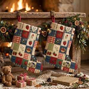 Op de afbeelding: Twee bijpassende kerstsokken met een patchworkontwerp van vierkanten met aardbeimotieven, bloemenpatronen en kleine huisillustraties in tinten rood, blauw, groen en crème, elk hangend aan een rode stoffen lus.