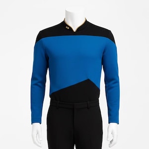 Könnte beinhalten: Ein langärmeliges Oberteil mit schwarzem Kragen und goldener Verzierung, einem blauen oberen Brustbereich und einem schwarzen unteren Teil mit kantigem Design. Dazu wird eine einfarbige schwarze Hose mit flacher Vorderseite kombiniert.