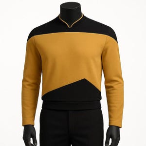 Puede incluir: Una camisa de manga larga con un diseño de bloques de color amarillo mostaza y negro. La prenda presenta un canesú superior negro y una sección inferior negra que se inclina hacia arriba, un cuello tipo mandarín con ribete amarillo.