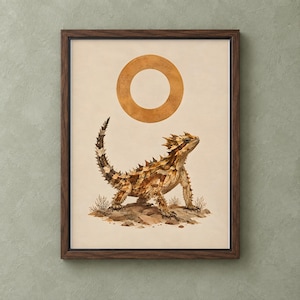 May include: A framed wall art print featuring a detailed illustration of a horned lizard standing on rocky terrain with small desert shrubs. Above the lizard is a large, solid orange circle. The frame is made of dark brown wood with a thin black border.