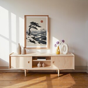May include: A framed wall art print featuring a landscape illustration of a lone, wind-swept pine tree on a snowy cliff overlooking a winding river. The scene includes distant mountains, a dark blue river, and a solid red sun against a cream background.