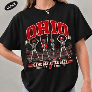 May include: A black t-shirt featuring a graphic of four skeletons in red and white football gear posing on a stadium field. The text reads OHIO at the top and GAME DAY AFTER DARK at the bottom, with a small red outline of the state of Ohio included.