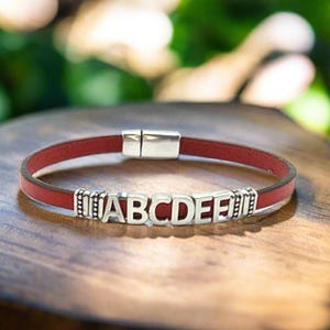 May include: A red leather bracelet with silver-colored letters spelling out ABCDEF. The bracelet has a magnetic clasp and is displayed on a wooden surface. The letters are in a bold, sans-serif font.