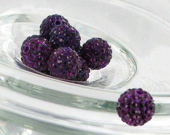 May include: Close-up of several dark purple, crystal-covered beads. The beads are round and have a textured surface, with a hole visible in each. They are displayed in a clear, glass dish, with one bead separate.