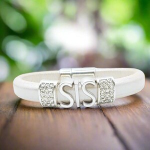 Könnte beinhalten: Weißes Lederarmband mit silberfarbenen Akzenten. Das Armband zeigt die Buchstaben "SIS" in der Mitte, flankiert von rechteckigen silbernen Verschlüssen, die mit kleinen, klaren Kristallen besetzt sind. Das Armband liegt auf einer Holzoberfläche.