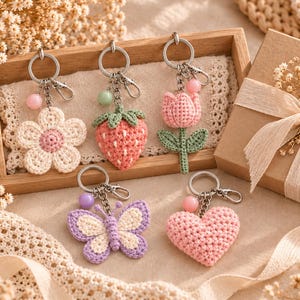May include: Five crochet keychains featuring a flower, a strawberry, a tulip, a butterfly, and a heart. Each keychain is attached to a silver metal ring with a small colored bead and a lobster clasp, showcasing intricate yarn textures and pastel colors.