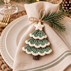 Handmade crochet Christmas tree ornament attached to a kraft paper wrapped holiday gift with twine, pine greenery, and rustic Christmas decorations.

Sage green crochet Christmas tree ornaments hanging from wooden wall hooks in a soft neutral nursery with cozy cream decor and wooden accents.

Rustic Christmas display featuring layered crochet tree ornaments in a woven basket beside pine branches, candles, pinecones, and warm holiday lights.
