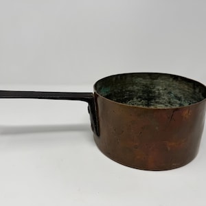 Puede incluir: Un cazo redondo de cobre con una pátina oscura y desgastada, con un mango largo, plano y de hierro negro unido al lateral. El interior del cazo muestra signos de oxidación y desgaste, con una superficie metálica texturizada en el exterior.