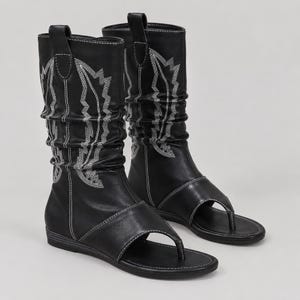 May include: A pair of black mid-calf boots featuring a thong-style open toe design. The boots have a slouchy shaft with white decorative stitching in a leaf-like pattern, a flat sole, and a pull tab at the top of each boot shaft.
