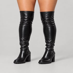 Puede incluir: Un par de botas negras por encima de la rodilla con acabado liso y mate, punta redondeada y tacón de bloque grueso. La caña de las botas se extiende por encima de la rodilla con una textura ligeramente fruncida y suela de goma dentada.