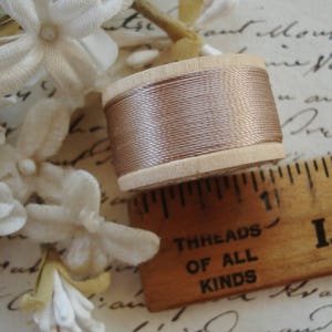 May include: A spool of light brown thread sits next to a wooden ruler with the text "THREADS OF ALL KINDS". The thread is wound around a wooden spool. White fabric flowers are in the background.