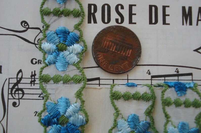 May include: White lace trim with blue and green embroidered flowers. The trim is shown next to a US penny.