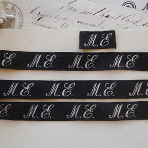 May include: Three black ribbon trims with white embroidered "M.E." lettering. The ribbon trims are arranged in a stack, with the top trim slightly offset to the right. A smaller black ribbon trim with white embroidered "M.E." lettering is positioned above the stack of trims.