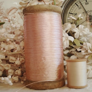May include: A large wooden spool of pale pink thread stands next to a smaller spool of cream-colored thread. The large spool features a weathered wooden base and top, while the smaller spool has a smooth, light-colored wooden core.