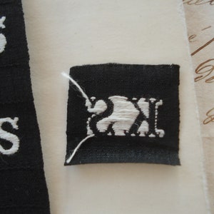 May include: A black fabric label with white embroidered letters "KS" and a white embroidered design of a stylized animal, possibly a bird or a fish, inside a square.