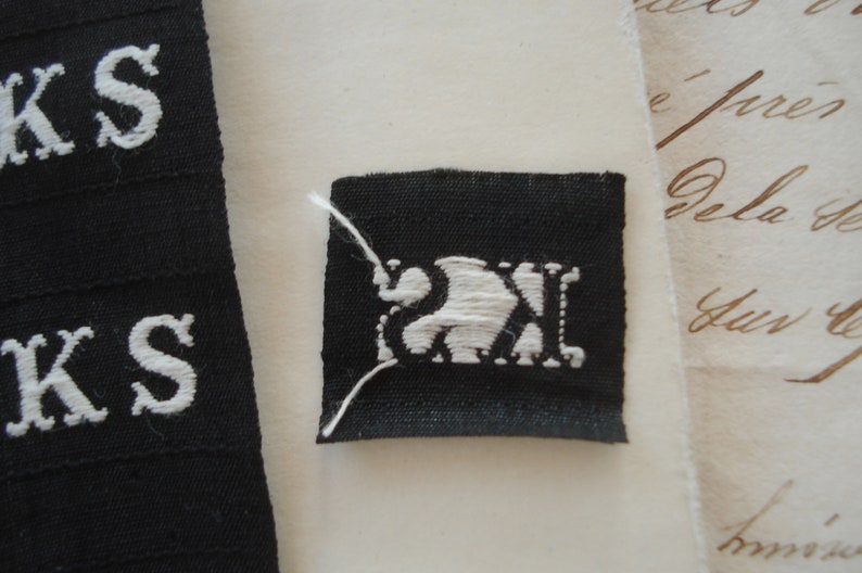 May include: A black fabric label with white embroidered letters "KS" and a white embroidered design of a stylized animal, possibly a bird or a fish, inside a square.