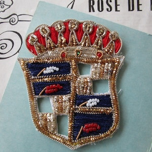 May include: A gold and silver embroidered crest with a red crown and blue and white sections. The crest is decorated with beads and sequins.