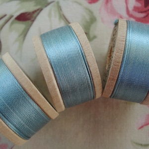May include: Three wooden spools of pale blue thread are arranged diagonally. The thread is tightly wound around the spools, and the wood has a natural, light brown colour. The background is a floral pattern.