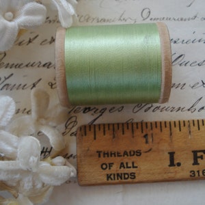 May include: A spool of light green thread sits on a vintage paper background, next to a wooden ruler. The ruler reads "THREADS OF ALL KINDS". Decorative white flowers and script are also in the image.