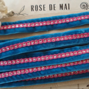 May include: Blue velvet ribbon with a pink and silver trim. The ribbon is folded in layers and the text "ROSE DE MAI" is visible in the background.