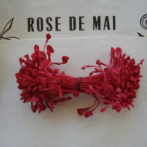 Può includere: Un mazzo di boccioli di fiori di seta rosso scuro legati insieme con uno spago marrone. I boccioli di fiori sono disposti a forma di fiocco. Il testo "ROSE DE MAI" è visibile sullo sfondo.