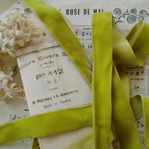 May include: A close-up of a spool of bright green velvet ribbon, with a vintage paper label and sheet music in the background. The label reads "9 METRES 15 GARANTIS MADE IN FRANCE". A cluster of white flowers is also visible.