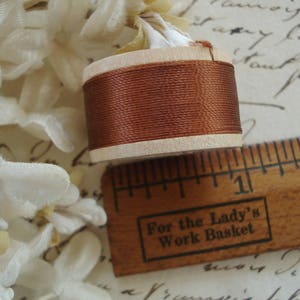 May include: A close-up of a wooden spool of brown thread, a wooden ruler, and white floral accents. The ruler has the text "For the Lady's Work Basket". The spool is approximately 1 inch in diameter.