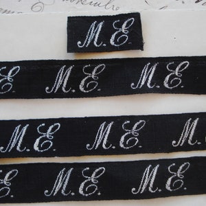 May include: Four strips of black fabric with white embroidered letters that read "M.E." and one small square of black fabric with white embroidered letters that read "M.E."