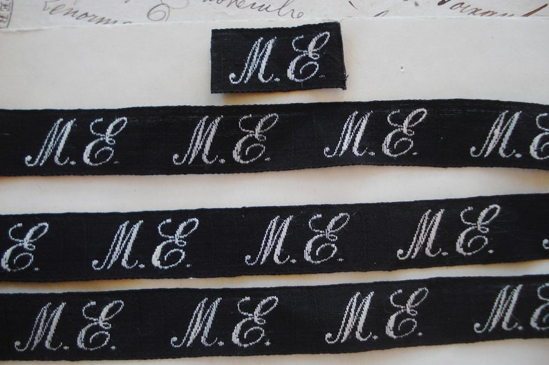 May include: Four strips of black fabric with white embroidered letters that read "M.E." and one small square of black fabric with white embroidered letters that read "M.E."