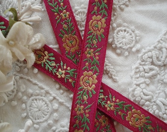 May include: Two pink satin ribbons with gold and green floral embroidery. The ribbons are crossed over each other.