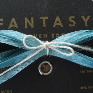 May include: A spool of blue ribbon with a white bow tied around it. The ribbon is labeled "Fantasy" and has a gold label with the text "Seven Edge 21% Rayon 20% Silk 100 Yds."