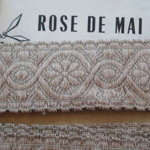 Puede incluir: Primer plano de un ribete de tela beige con un patr&oacute;n repetitivo de flores tejidas. El ribete tiene un borde deshilachado y est&aacute; etiquetado como "ROSE DE MAI".