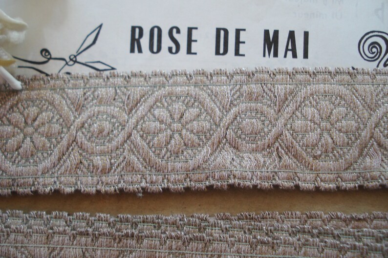 Puede incluir: Primer plano de un ribete de tela beige con un patr&oacute;n repetitivo de flores tejidas. El ribete tiene un borde deshilachado y est&aacute; etiquetado como "ROSE DE MAI".