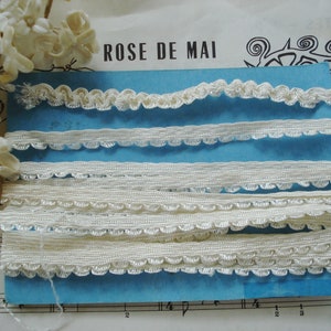 May include: Five spools of white lace trim with scalloped edges. The trim is arranged on a blue background with the text "ROSE DE MAI" above it.