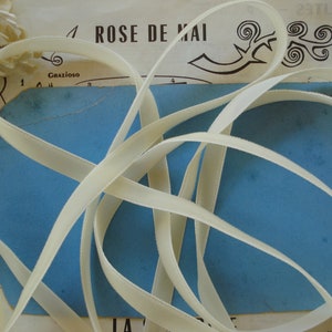 Könnte beinhalten: Nahaufnahme einer Spule mit cremefarbenem Satinband auf einer blauen Oberfläche. Das Band ist in einem dekorativen Muster angeordnet. Der Text "ROSE DE MAI" ist im Hintergrund sichtbar.