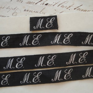 May include: Black and white fabric ribbon with the initials "M.E." repeated multiple times. The ribbon is approximately 1 inch wide.