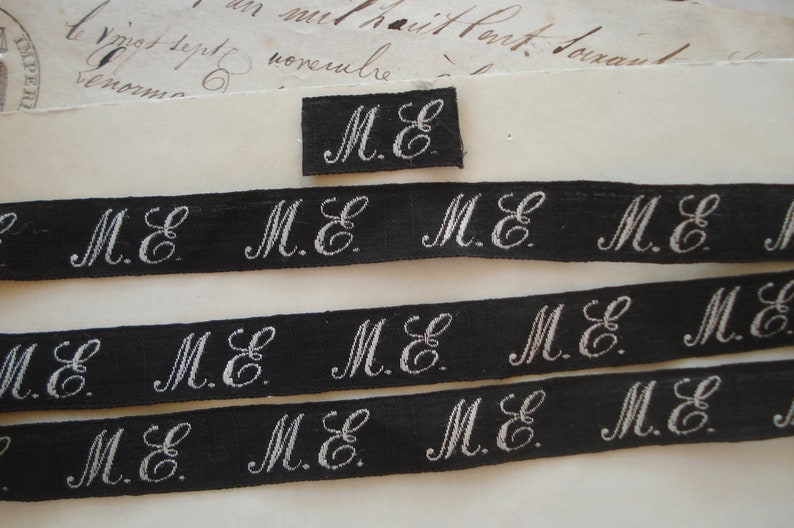 May include: Black and white fabric ribbon with the initials "M.E." repeated multiple times. The ribbon is approximately 1 inch wide.