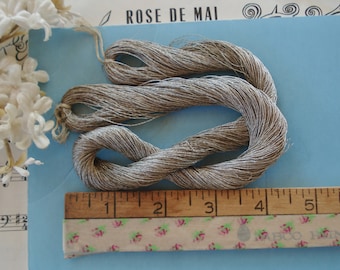 May include: A coiled bundle of metallic silver embroidery thread positioned above a wooden ruler that shows the thread measures approximately 5.5 inches in length. The thread has a shimmering, reflective texture and is composed of many fine strands.