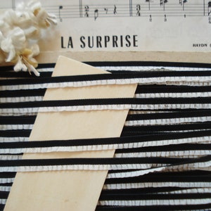 May include: Black and white striped ribbon with a ruffled edge. The ribbon is folded and stacked on top of a piece of wood. The text "LA SURPRISE" is visible in the background.