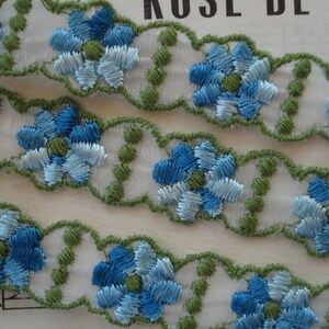 May include: White lace trim with blue and green embroidered flowers. The trim has a scalloped edge.