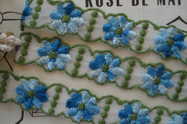 May include: White lace trim with blue and green embroidered flowers. The trim has a scalloped edge.