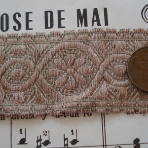 Puede incluir: Primer plano de un ribete de tela marr&oacute;n con un patr&oacute;n geom&eacute;trico repetitivo. El ribete est&aacute; sobre una hoja de m&uacute;sica con las palabras "ROSE DE MAI" impresas en la parte superior. Una moneda de un centavo estadounidense de color cobre est&aacute; sobre el ribete.