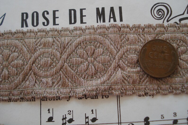 Puede incluir: Primer plano de un ribete de tela marr&oacute;n con un patr&oacute;n geom&eacute;trico repetitivo. El ribete est&aacute; sobre una hoja de m&uacute;sica con las palabras "ROSE DE MAI" impresas en la parte superior. Una moneda de un centavo estadounidense de color cobre est&aacute; sobre el ribete.