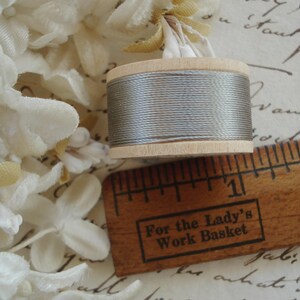 May include: A wooden spool of grey thread, a vintage ruler with the text "For the Lady's Work Basket", and white fabric flowers are arranged on a surface with handwritten script. The thread is wound around a wooden spool.