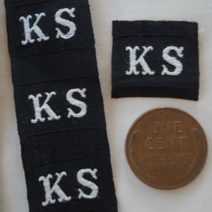 May include: Close-up of five black fabric labels with white embroidered letters spelling "KS". A single copper penny is also visible in the image.